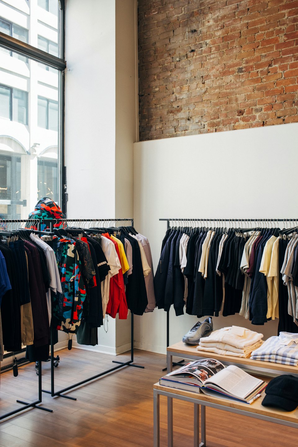 Clothing boutique displays apparel on racks and tables with brick wall.
