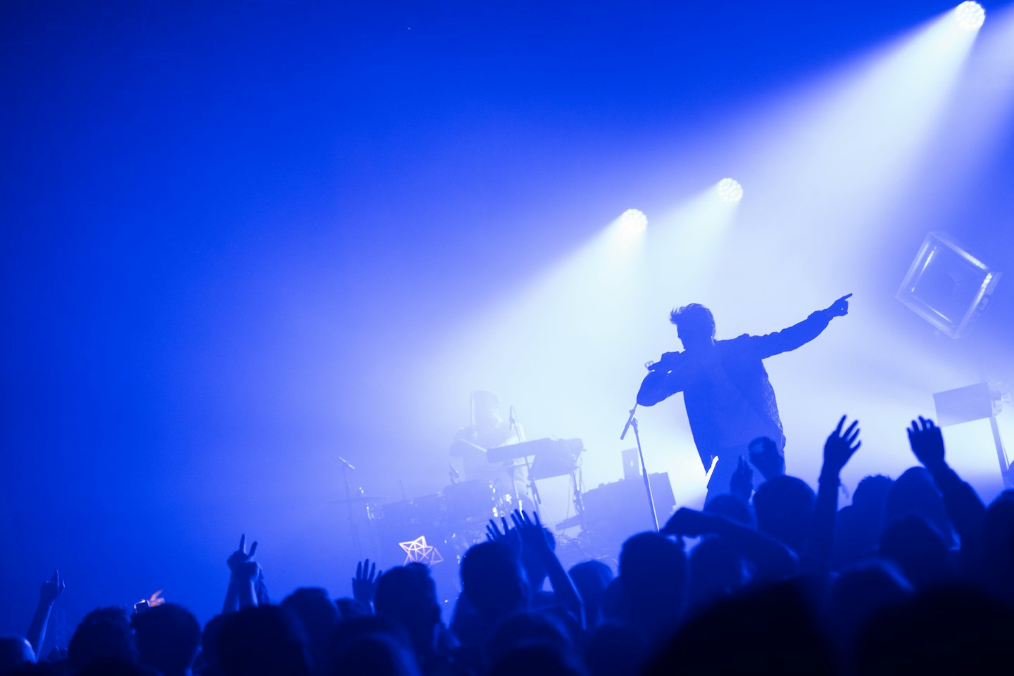 Singer in silhouette on stage with blue lights and crowd hands up.